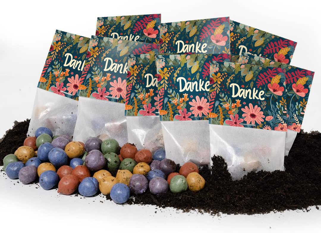 Mehrere bunte Tütchen mit der Aufschrift "Danke" und Blumenmuster stehen auf Erde, davor liegen bunte Samenbälle.