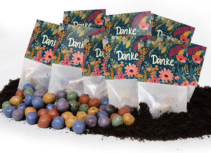 Mehrere bunte Tütchen mit der Aufschrift "Danke" und Blumenmuster stehen auf Erde, davor liegen bunte Samenbälle.