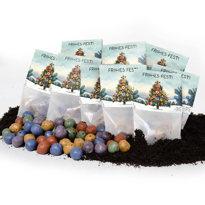 Mehrere Tütchen mit der Aufschrift "Frohes Fest!" und einem weihnachtlich geschmückten Baum, davor liegen bunte Samenbälle auf Erde.