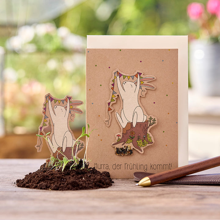 Grußkarte mit Illustration eines fröhlichen Hasen, der Wimpel hält, auf einem Baumstumpf; Text: "Hurra, der Frühling kommt!", im Vordergrund kleine Pflänzchen in Erde.