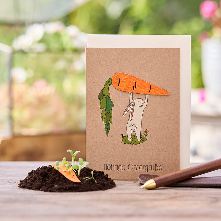 Osterkarte mit gezeichnetem Hasen, der eine große Karotte hält, daneben Erde mit kleinen Pflanzen und einer Karottenverzierung, Text „Möhrige Ostergrüße!“.