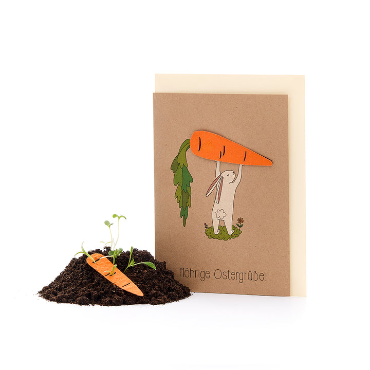 Eine Osterkarte mit einem gezeichneten Hasen, der eine große Karotte über dem Kopf hält, daneben ein kleiner Erdhaufen mit einer Papier-Karotte und keimenden Pflänzchen. Text: "Möhrige Ostergrüße!"