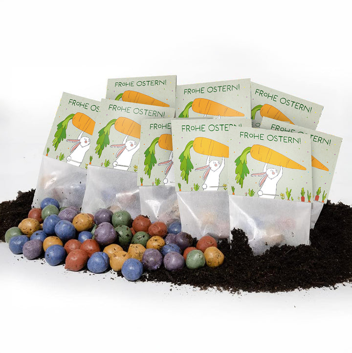 Mehrere Ostern-Samentütchen mit der Aufschrift "Frohe Ostern!" und bunter Samen-Konfetti auf Erde.