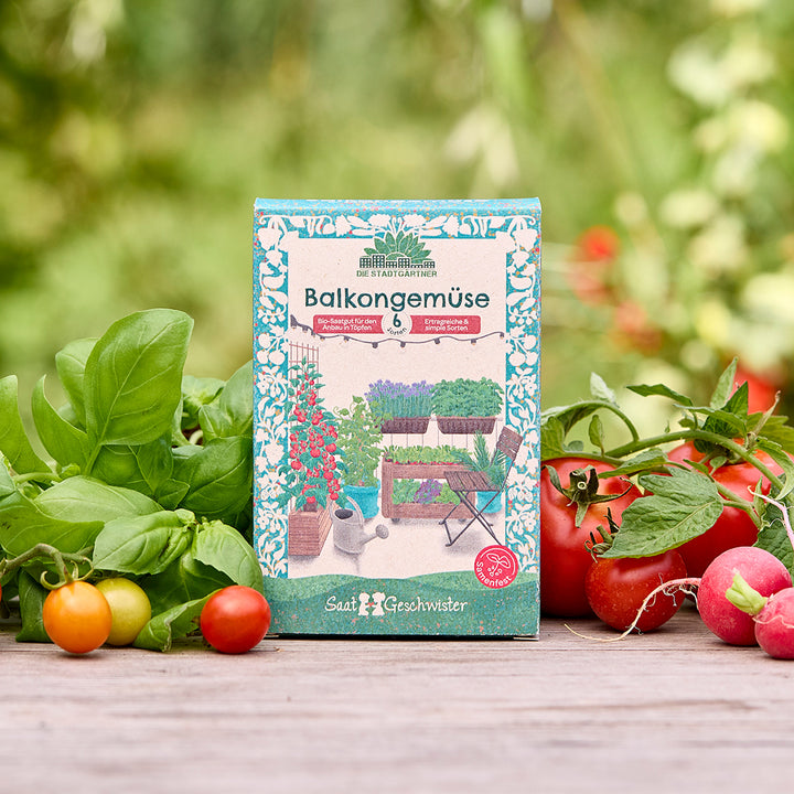 Paket "Balkongemüse" mit Samen für verschiedene Gemüsesorten auf einem Holztisch, umgeben von frischen Kräutern, Tomaten und Radieschen im Freien.
