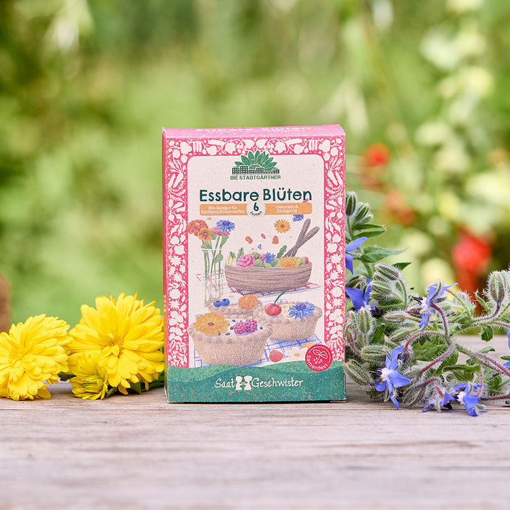 Samenpackung mit der Aufschrift "Essbare Blüten" steht auf einem Holztisch, umgeben von gelben und lilafarbenen Blüten, unscharfer grüner Hintergrund.