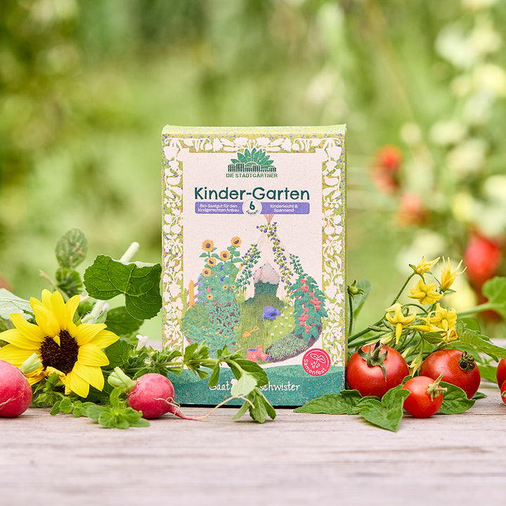 Eine Packung „Kinder-Garten“ Bio-Saatgut steht auf einem Holztisch, umgeben von Sonnenblume, Radieschen, Tomaten und frischem Grün im Garten.