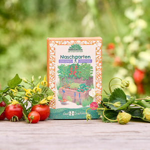 Eine Packung "Naschgarten" Saatgut steht auf einem Holztisch im Garten, umgeben von frischen Tomaten, Erdbeeren und grünem Laub.