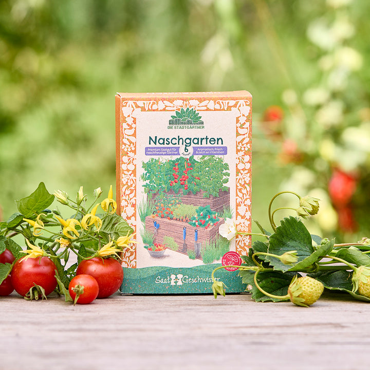 Eine Packung "Naschgarten" Saatgut steht auf einem Holztisch im Garten, umgeben von frischen Tomaten, Erdbeeren und grünem Laub.