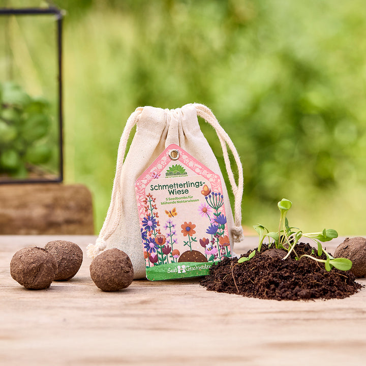 Stoffsäckchen mit der Aufschrift „Schmetterlings-Wiese“, daneben Seedballs und Erde mit jungen Pflanzen, im Hintergrund unscharfe Natur.