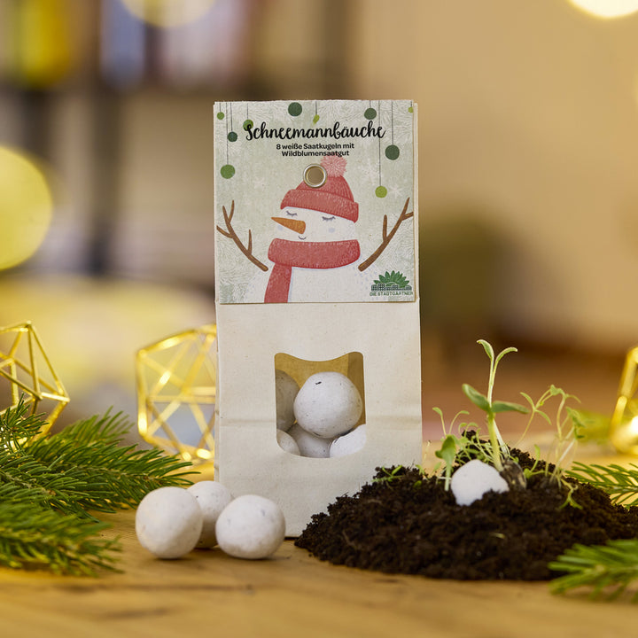 Eine Packung mit weißen Saatkugeln namens "Schneemannbäuche" steht auf einem Tisch, daneben Erde, Setzlinge und Tannenzweige.