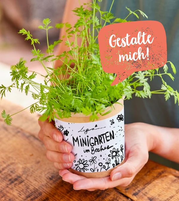 Person hält einen Becher mit Kräuterpflanze, auf dem "Minigarten im Becher" und "Gestalte mich!" steht.