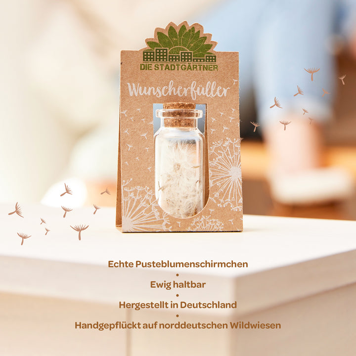 Wunschflasche mit Pusteblumenschirmchen in einem Glasfläschchen, umgeben von einer bedruckten Papierverpackung mit der Aufschrift 'Die Stadtgärtner' und 'Wunscherfüller', stehend auf einer weißen Oberfläche mit unscharfem Hintergrund."
