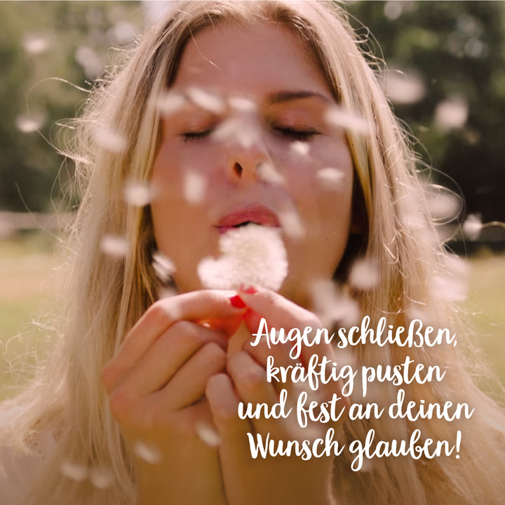 Frau bläst Pusteblume mit geschlossenen Augen und zerzausten Samen in der Luft, Text: "Augen schließen, kräftig pusten und fest an deinen Wunsch glauben!"