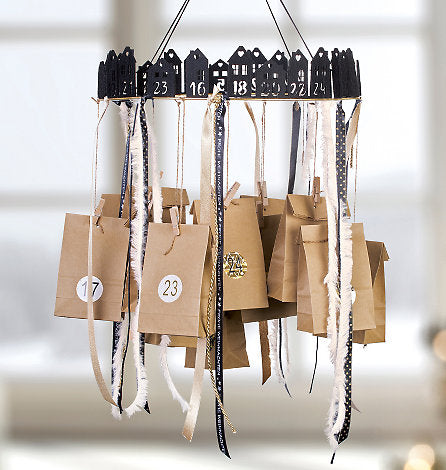 Selbstgemachter Adventskalender aus braunen Papiertüten, die an einem runden Gestell mit schwarzen Häusern und Bändern hängen.