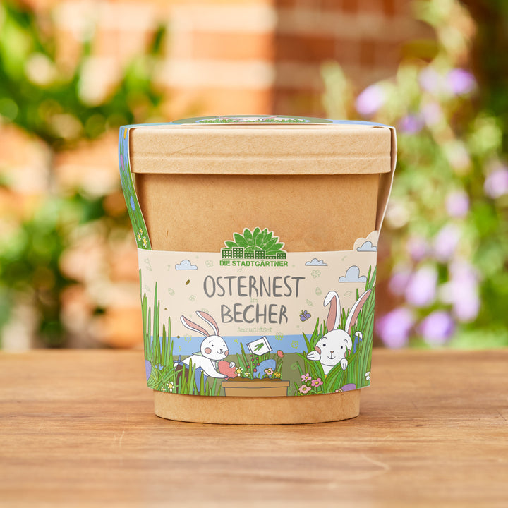 Pappbecher mit Osterdesign und der Aufschrift „Osternest im Becher“ auf einem Holztisch, im Hintergrund unscharfe Pflanzen.