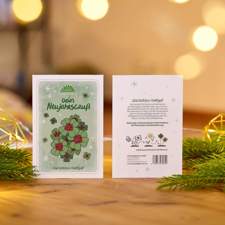 Zwei Neujahrskarten mit Glücksklee-Motiv und Text auf einem Holztisch, dekoriert mit Tannenzweigen und Lichtern im Hintergrund.