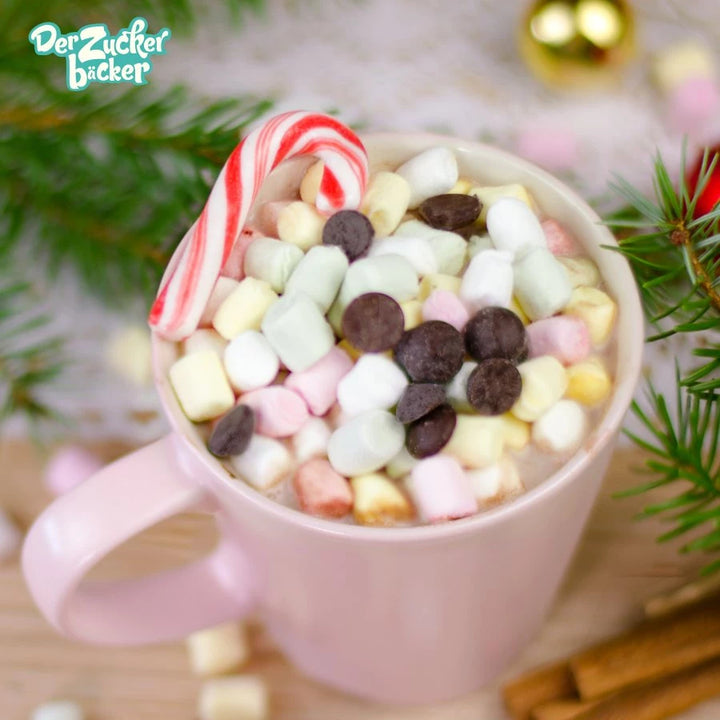 Eine Tasse heiße Schokolade mit Mini-Marshmallows und Schokostückchen, daneben ein Zuckerstangen, umgeben von Tannenzweigen und Weihnachtsdekoration.