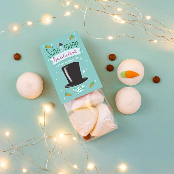 Verpackung mit "Schneemann Bausatz" Marshmallows, umgeben von kleinen Schokoladenkugeln und Lichterketten auf mintgrünem Hintergrund.