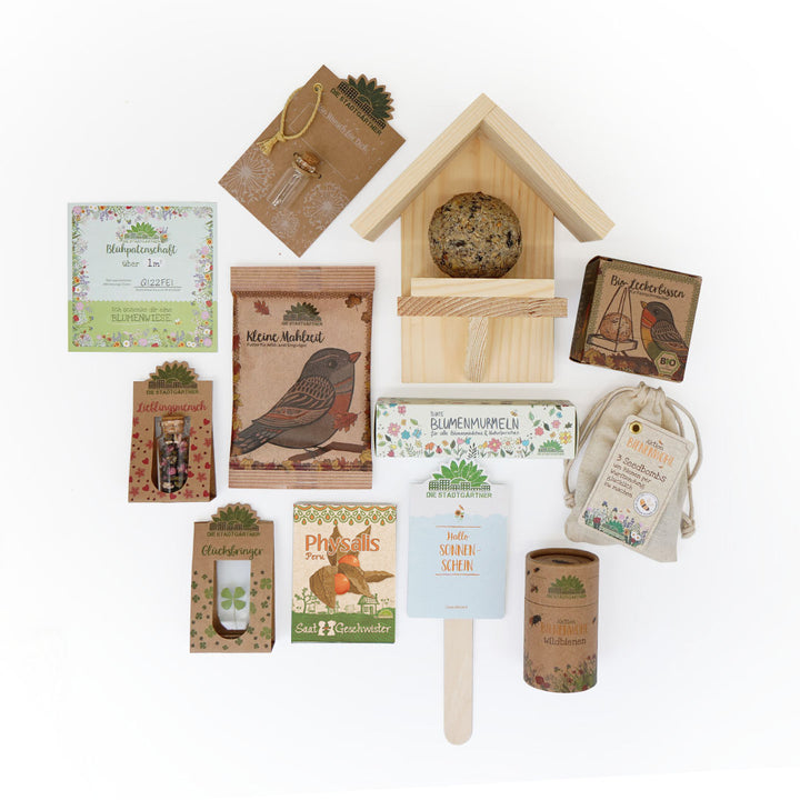 Verschiedene umweltfreundliche Garten- und Naturprodukte, darunter Saatgut, Pflanzenzubehör, ein Vogelhaus mit Futter sowie kleine Geschenkartikel, auf weißem Hintergrund arrangiert.