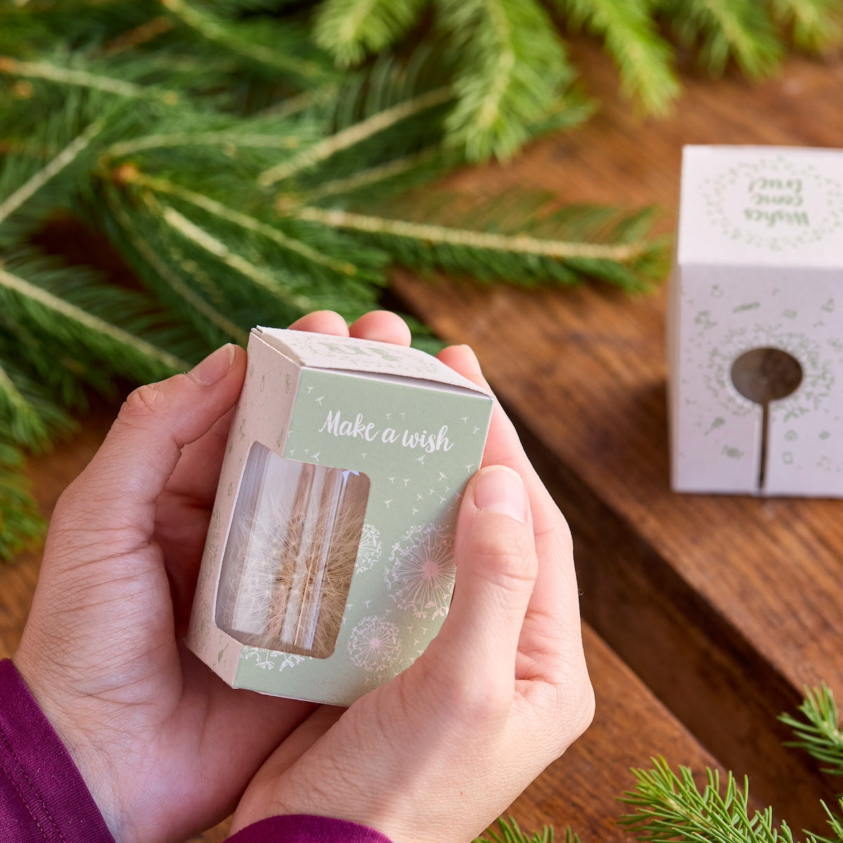 Hände halten eine kleine Geschenkbox mit der Aufschrift "Make a wish" neben Tannenzweigen auf einem Holztisch.