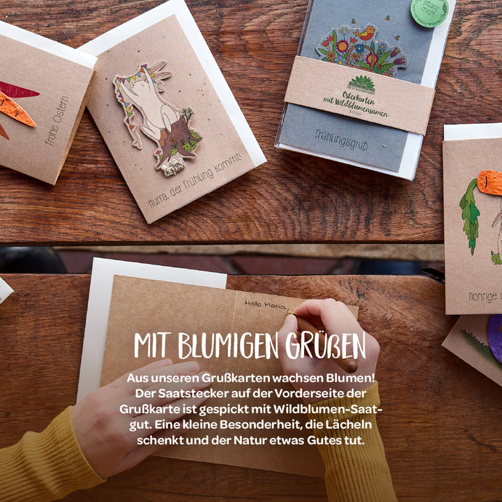 Mehrere bunte Grußkarten mit Motiven und Texten zu Frühling und Ostern liegen auf einem Holztisch; eine Person schreibt eine Karte.