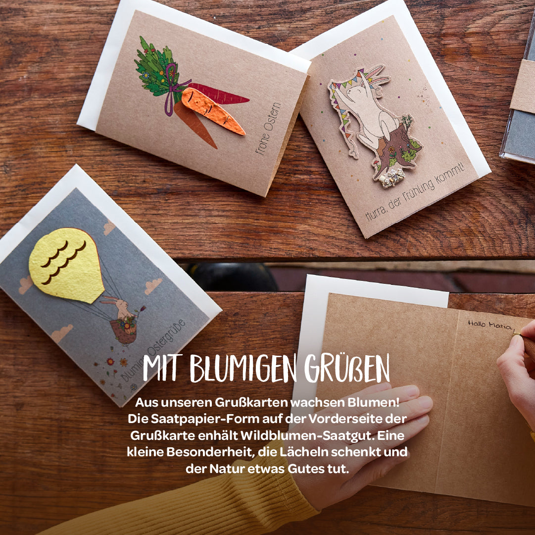 Vier verschiedene Grußkarten aus umweltfreundlichem Papier mit aufgeklebten Motiven aus Saatpapier, auf einem Holztisch liegend, darunter eine Person, die eine Karte öffnet.