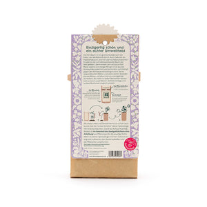 Rückseite einer Verpackung mit lila Blumenmuster, Text zur Umweltfreundlichkeit des Kiribaums, Anleitung zum Pflanzen, und Samenfest-Logo unten rechts.