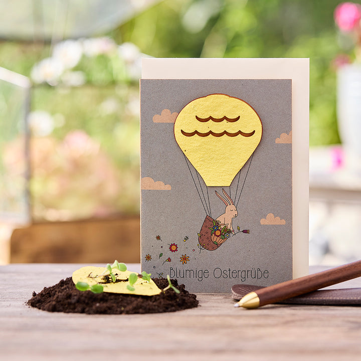 Osterkarte mit Hase im Heißluftballon, daneben Erde mit keimenden Pflänzchen und ein Stift.