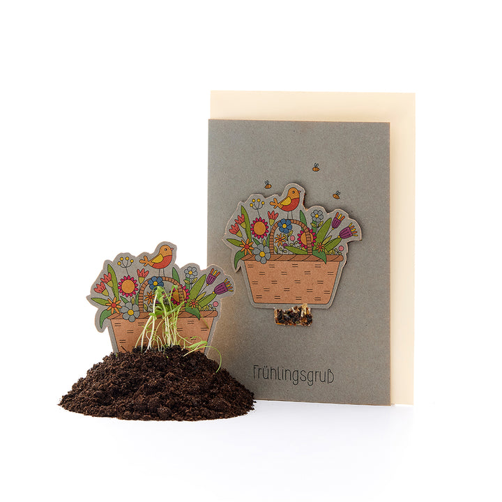 Grußkarte mit Blumenmotiv und Vogel, dazu ein Blumenkörbchen aus Papier im Erdhaufen mit sprießenden Pflänzchen, Text „Frühlingsgruß“.