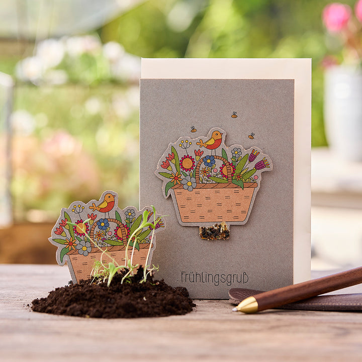 Grußkarte mit Frühlingsmotiv, Blumenkorb und Vogel, daneben eine Erde mit kleinen Setzlingen und Stift auf Holztisch.