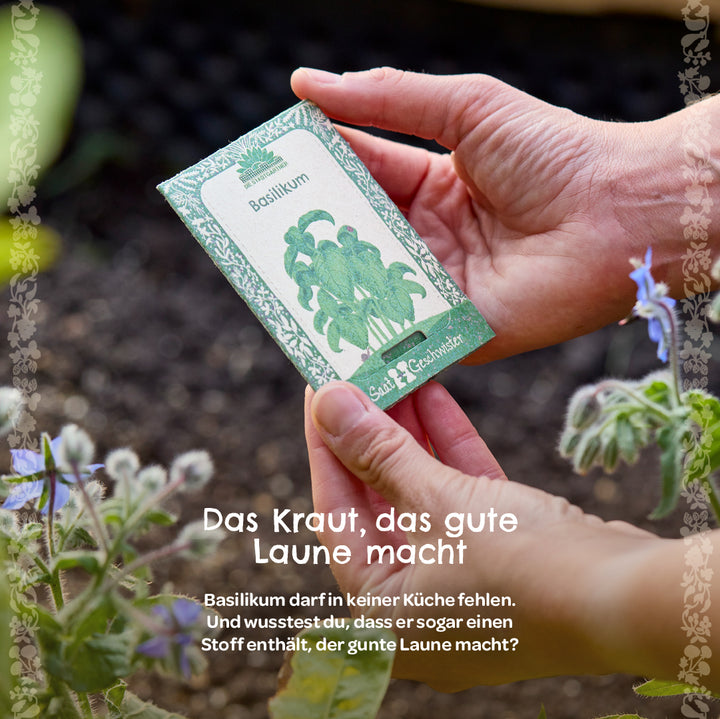 Eine Person hält eine Tüte mit Basilikumsamen im Garten, im Hintergrund sind Pflanzen und Erde zu sehen.