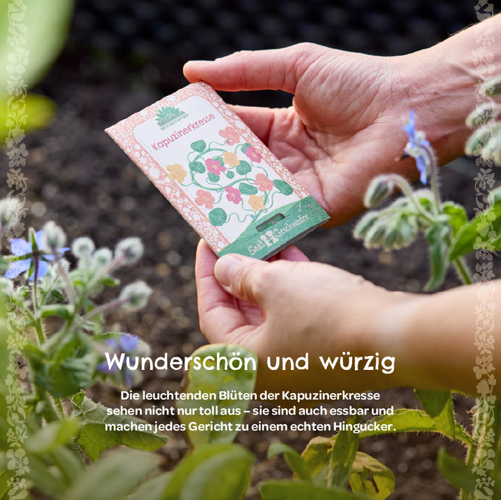 Hände halten ein Samentütchen mit der Aufschrift „Kapuzinerkresse“ im Garten, daneben blühende Pflanzen.