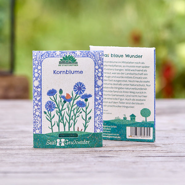 Zwei Samentütchen für Kornblumen der Marke "Saatgeschwister" auf einem Holztisch, mit floraler Illustration und Produktbeschreibung, im Hintergrund unscharfe Natur.