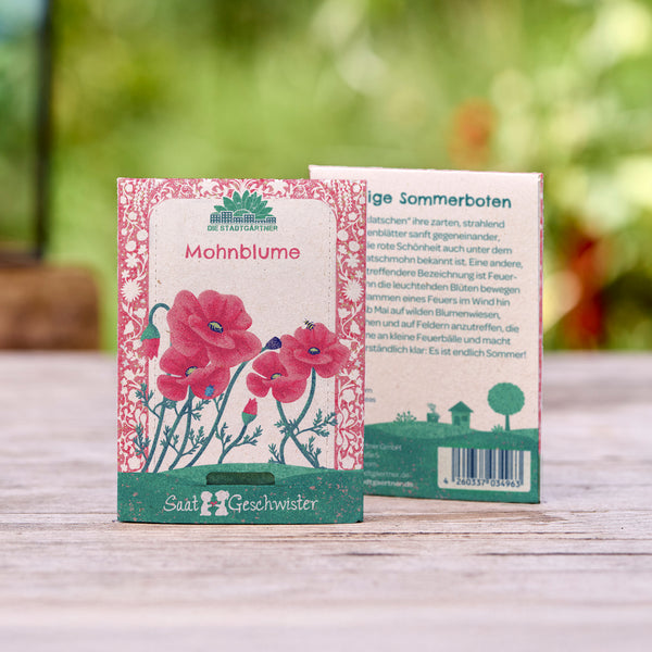 Zwei Samenpäckchen für Mohnblumen mit illustrierten roten Blumen auf Holzuntergrund und unscharfem Gartenhintergrund.