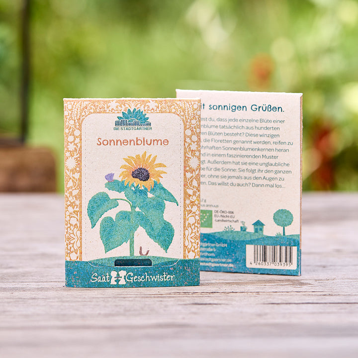 Zwei Saatgutpackungen für Sonnenblumen der Marke "Die Stadtgärtner" auf einem Holztisch, Vorder- und Rückseite sichtbar, mit Illustration einer Sonnenblume.
