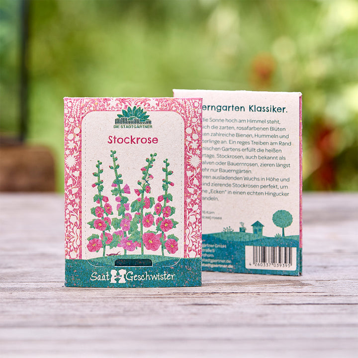 Zwei Saatgutpackungen für Stockrosen mit farbenfrohem Design und Text auf Deutsch, im Freien auf einem Holztisch platziert.