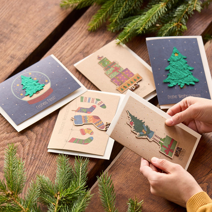 Mehrere weihnachtliche Grußkarten mit bunten Motiven und Tannenzweigen auf einem Holztisch, eine Karte wird von Händen gehalten.
