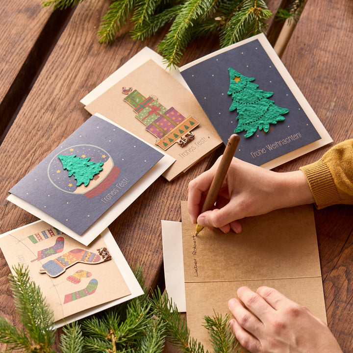 Eine Person schreibt eine Weihnachtskarte auf einem Holztisch, umgeben von weiteren festlichen Grußkarten und Tannenzweigen.