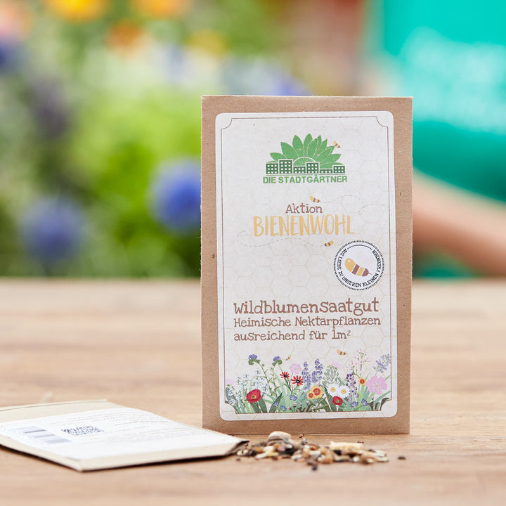 Papiertüte mit Wildblumensaatgut „Aktion Bienenwohl“ auf einem Holztisch, im Hintergrund unscharfe bunte Blumen.