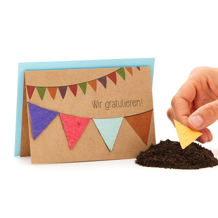 Glückwunschkarte mit bunter Wimpelkette und Schriftzug "Wir gratulieren!", Hand hält ein gelbes Dreieck über einem Erdhaufen.