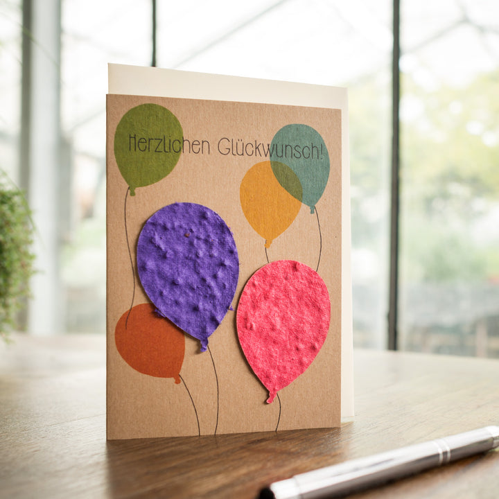 Glückwunschkarte mit bunten Ballons und Schriftzug "Herzlichen Glückwunsch!" auf einem Holztisch.