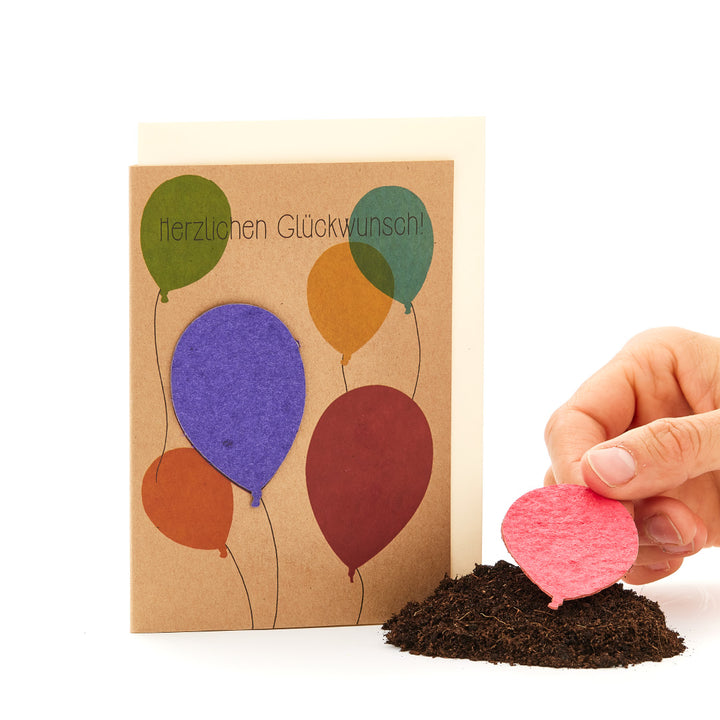 Glückwunschkarte mit bunten Ballons und der Aufschrift "Herzlichen Glückwunsch", neben einem Haufen Erde und einer Hand, die einen rosafarbenen Ballon pflanzt.