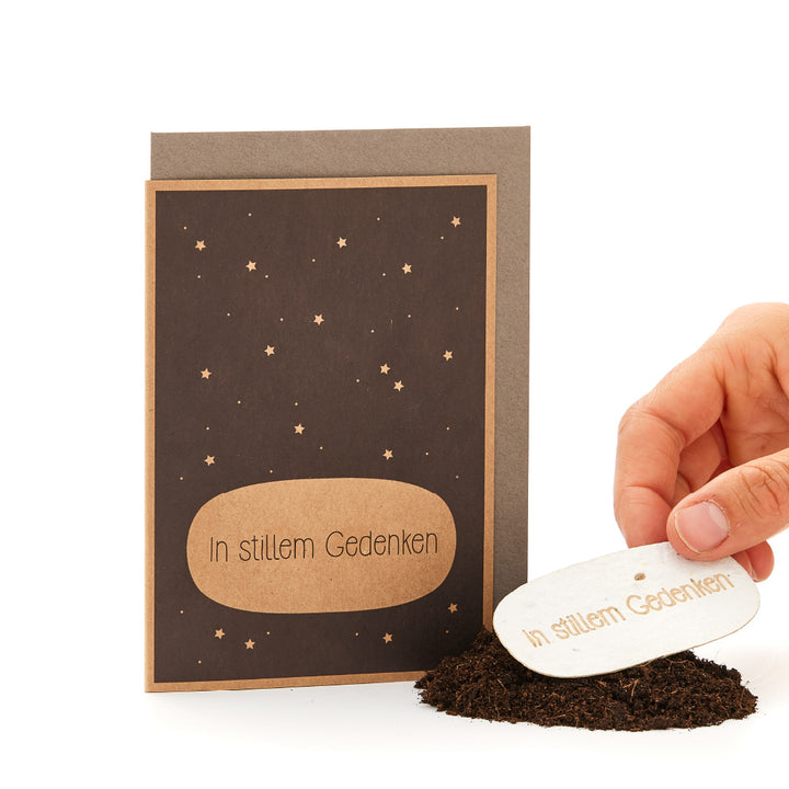 Eine Beileidskarte mit der Aufschrift "In stillem Gedenken" neben einer Hand, die ein oval geformtes Schild mit derselben Aufschrift über ein Häufchen Erde hält.