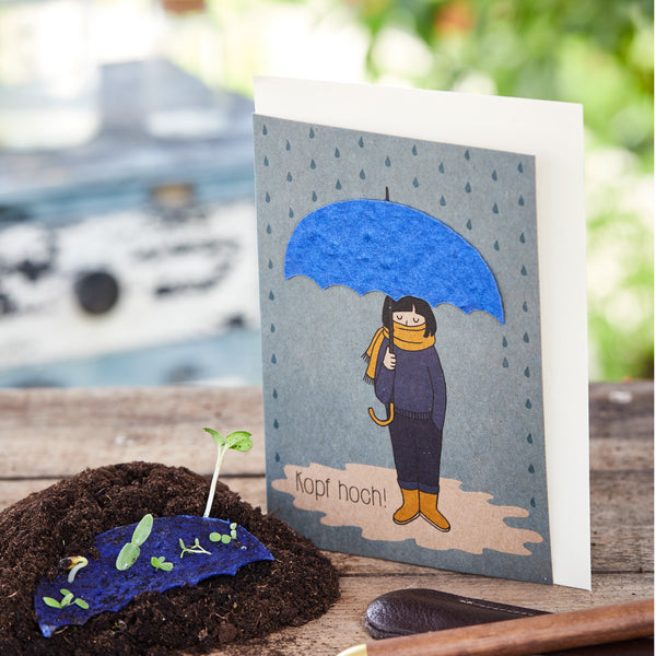 Grußkarte mit Zeichnung einer Person unter einem blauen Regenschirm und der Aufschrift "Kopf hoch!", neben einer jungen Pflanze auf Erde.