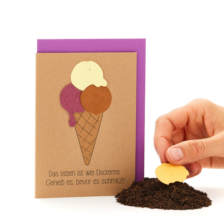 Hand, die ein herzförmiges Keks in Erdboden steckt, neben einer Grußkarte mit Eistüten-Motiv und dem Spruch 'Das Leben ist wie Eiscreme. Genieß es, bevor es schmilzt!'"