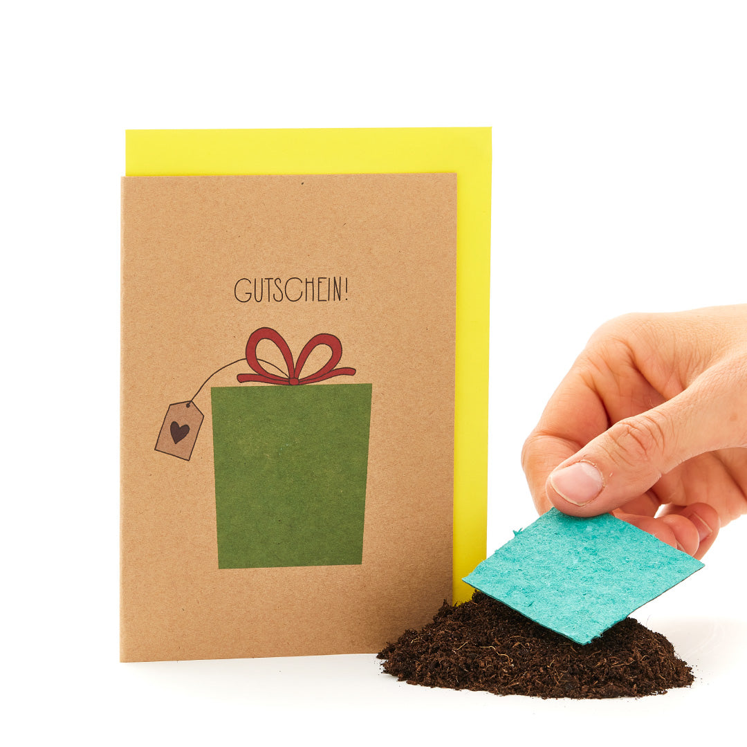 Hand hält eine Grußkarte mit der Aufschrift "GUTSCHEIN" und einem gezeichneten Geschenk, neben einem Haufen Erde mit einem Stück blauer Pappe.