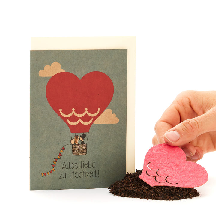 Glückwunschkarte zur Hochzeit mit Motiv eines Heißluftballons in Herzform und Hand, die ein Papierherz in Erde pflanzt.