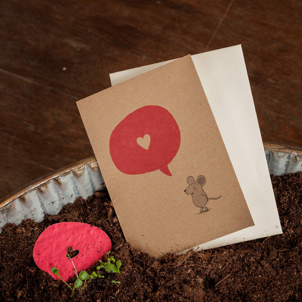Eine Grußkarte mit einer gezeichneten Maus und einer roten Sprechblase mit Herz, daneben ein weißer Umschlag und ein rotes Herz aus Papier in Erde.