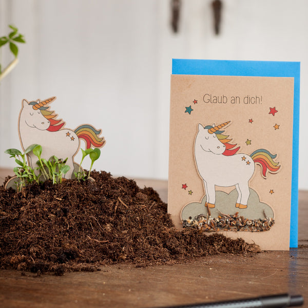 Ein Grußkarte mit einem Einhorn und dem Text "Glaub an dich!" neben einem kleinen Einhorn-Ausschnitt, umgeben von Erde.