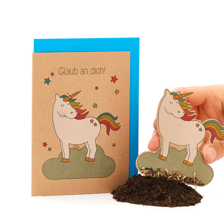 Grußkarte mit Einhorn und Regenbogen, Text "Glaub an dich!", daneben eine Hand, die ein passendes Einhorn-Papier in Erde steckt.